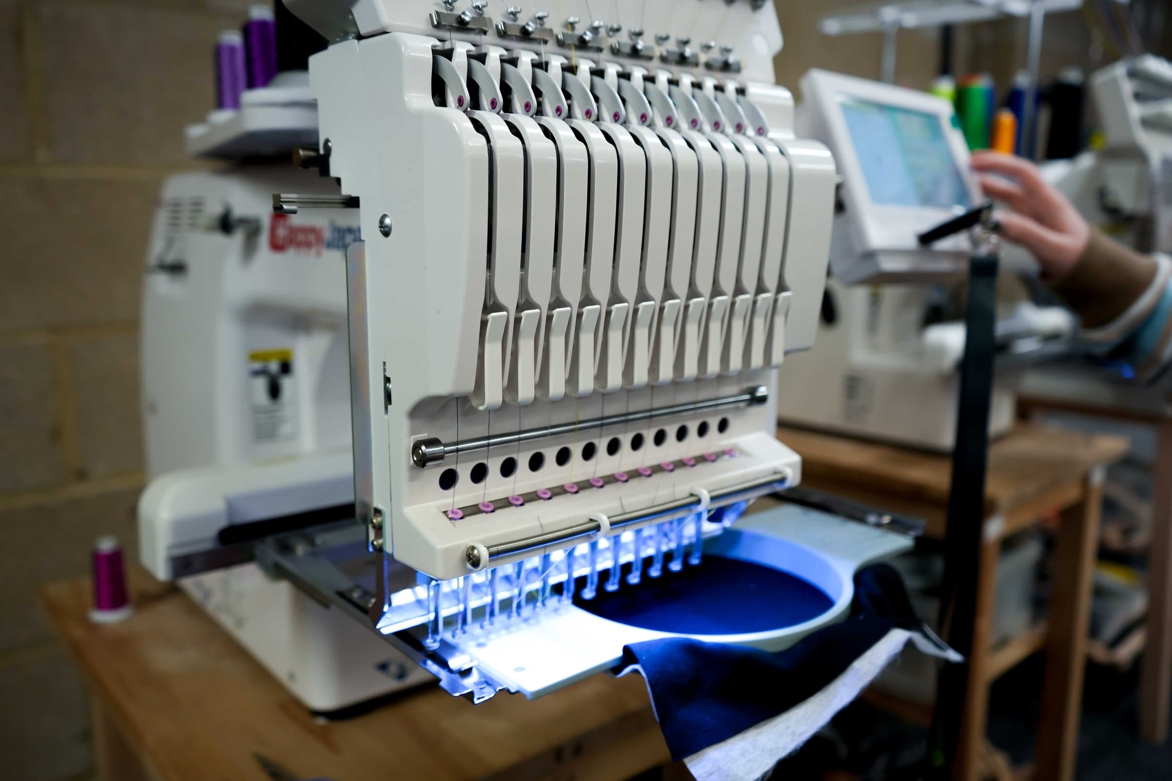 Multi-needle embroidery machine part-way through a run on navy fabric, with an operator setting the design on the touchscreen
